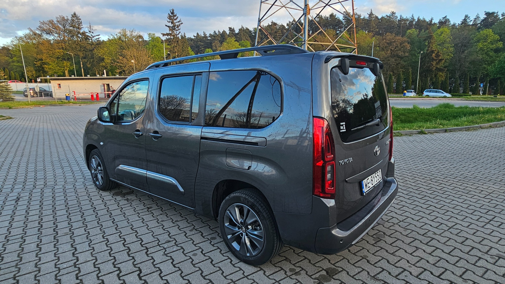 Szary van Toyota Proace City Verso z relingami dachowymi, zaparkowany na kostce brukowej, widok z tyłu i boku, z widoczną naklejką 'Toyota Nr 1' na tylnej szybie.