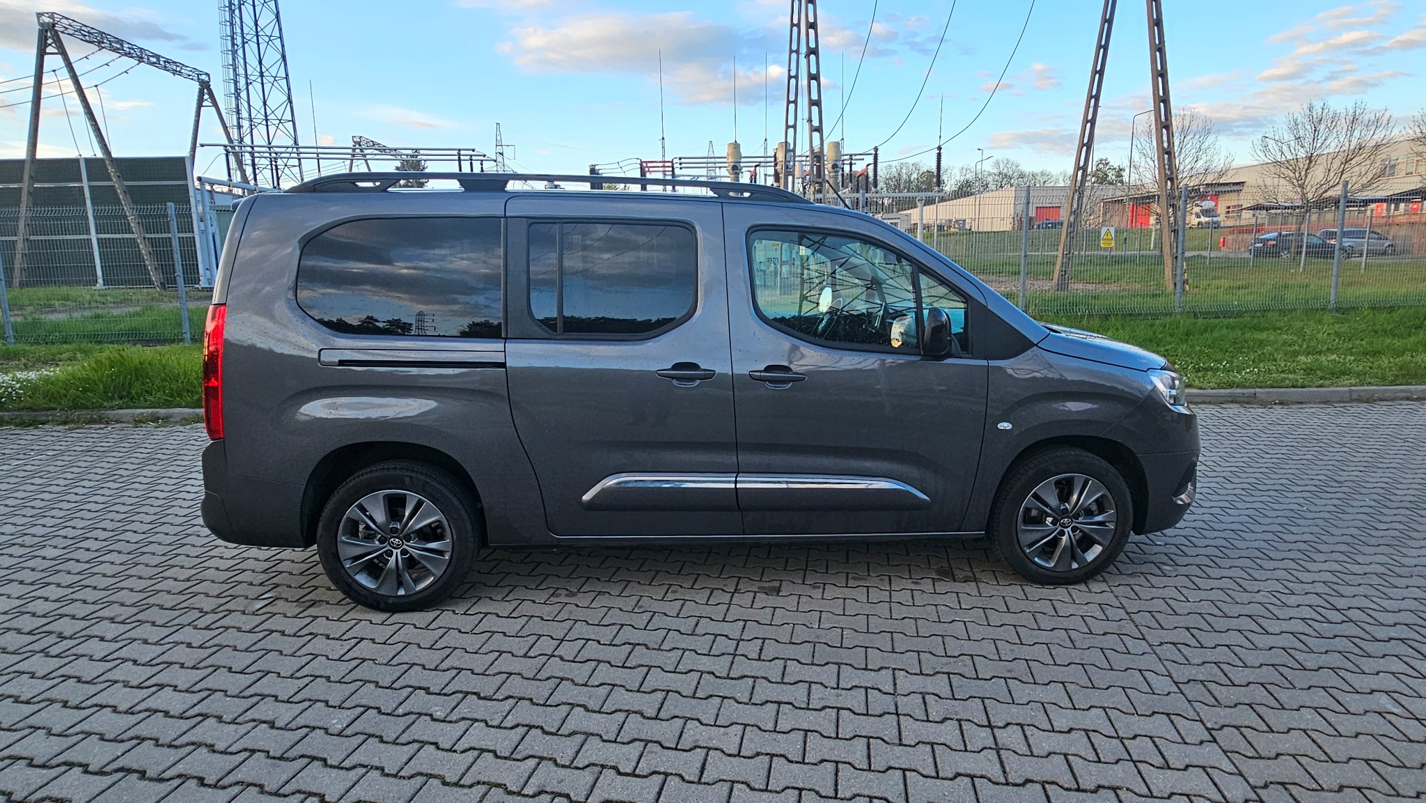 Szary van z relingami dachowymi, zaparkowany na kostce brukowej, z widoczną w tle infrastrukturą elektryczną i budynkiem.