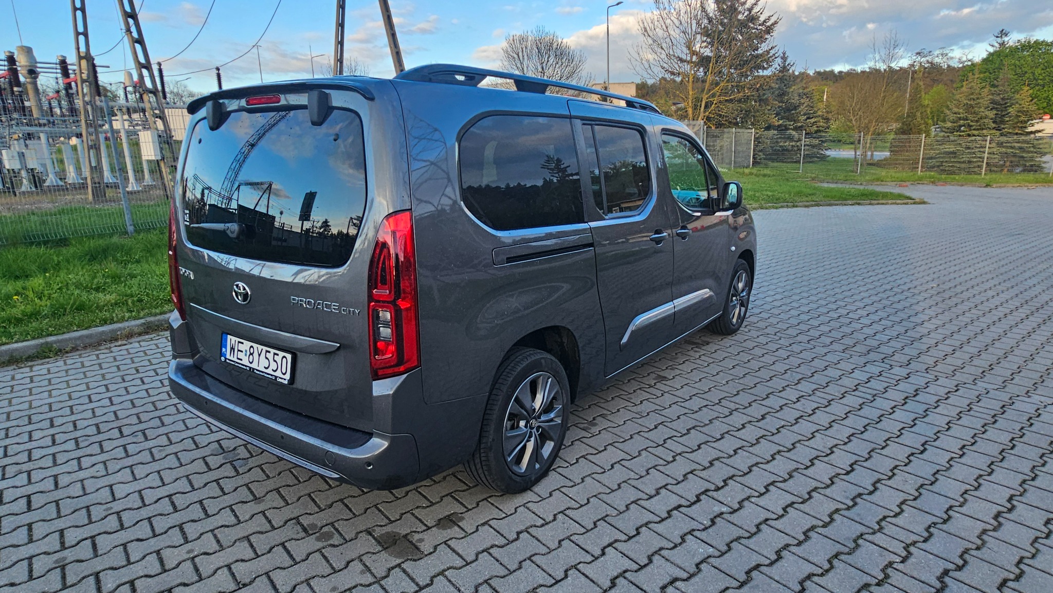 Szary samochód Toyota Proace City z relingami dachowymi, zaparkowany na kostce brukowej, z widoczną tablicą rejestracyjną i odbiciem konstrukcji w szybie.