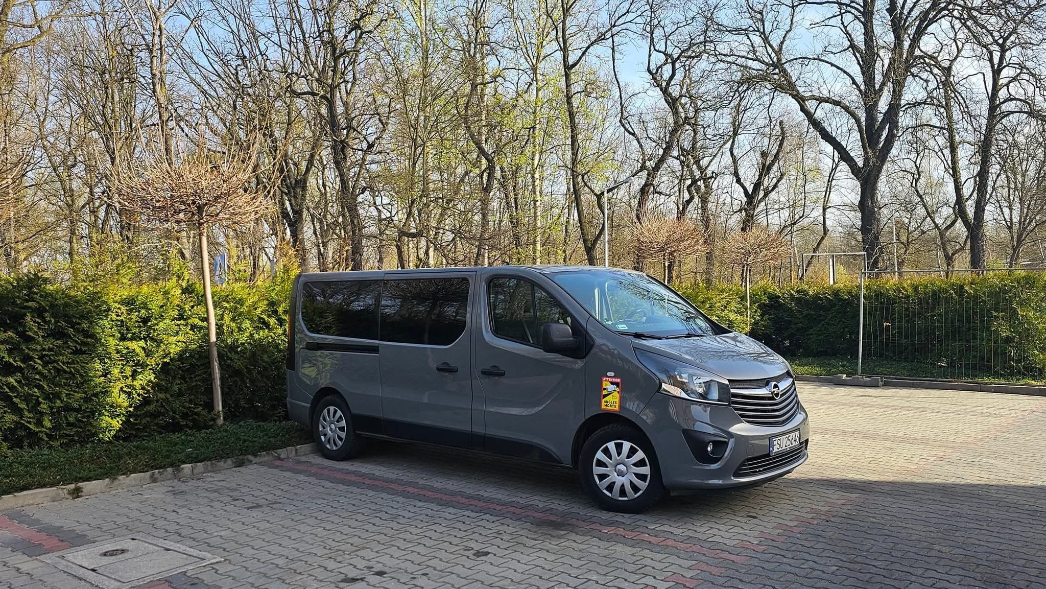 Szary Opel Vivaro z naklejką ostrzegawczą na parkingu, otoczony zielenią i drzewami. Widoczny numer rejestracyjny pojazdu. Dzień.