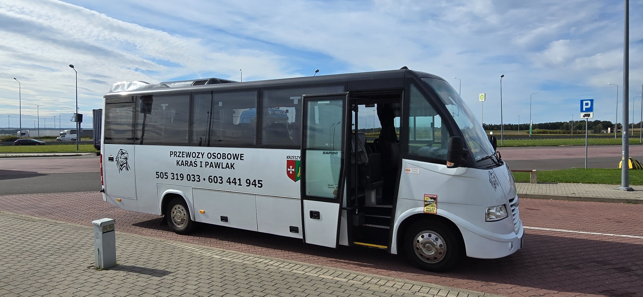 Biały bus z otwartymi drzwiami na parkingu, z napisem 'Przewozy Osobowe Karaś i Pawlak' oraz numerami telefonów. Widoczny znak parkingowy w tle.