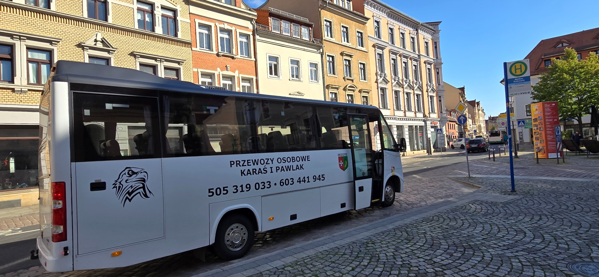 Biały bus z otwartymi drzwiami na tle miejskiej zabudowy. Na pojeździe logo firmy 'Przewozy Osobowe Karaś i Pawlak' oraz numer telefonu. Kostka brukowa na pierwszym planie.