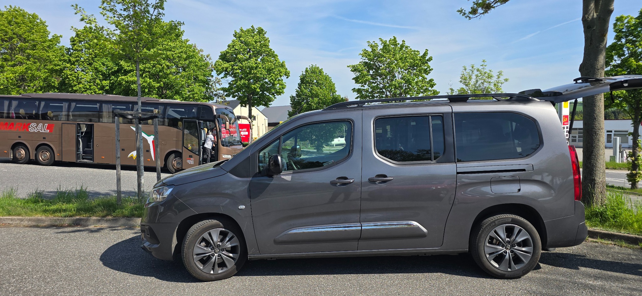 Szary samochód osobowy typu van z otwartą klapą bagażnika, na parkingu, w tle brązowy autobus z napisem MARKSAL i zielone drzewa.