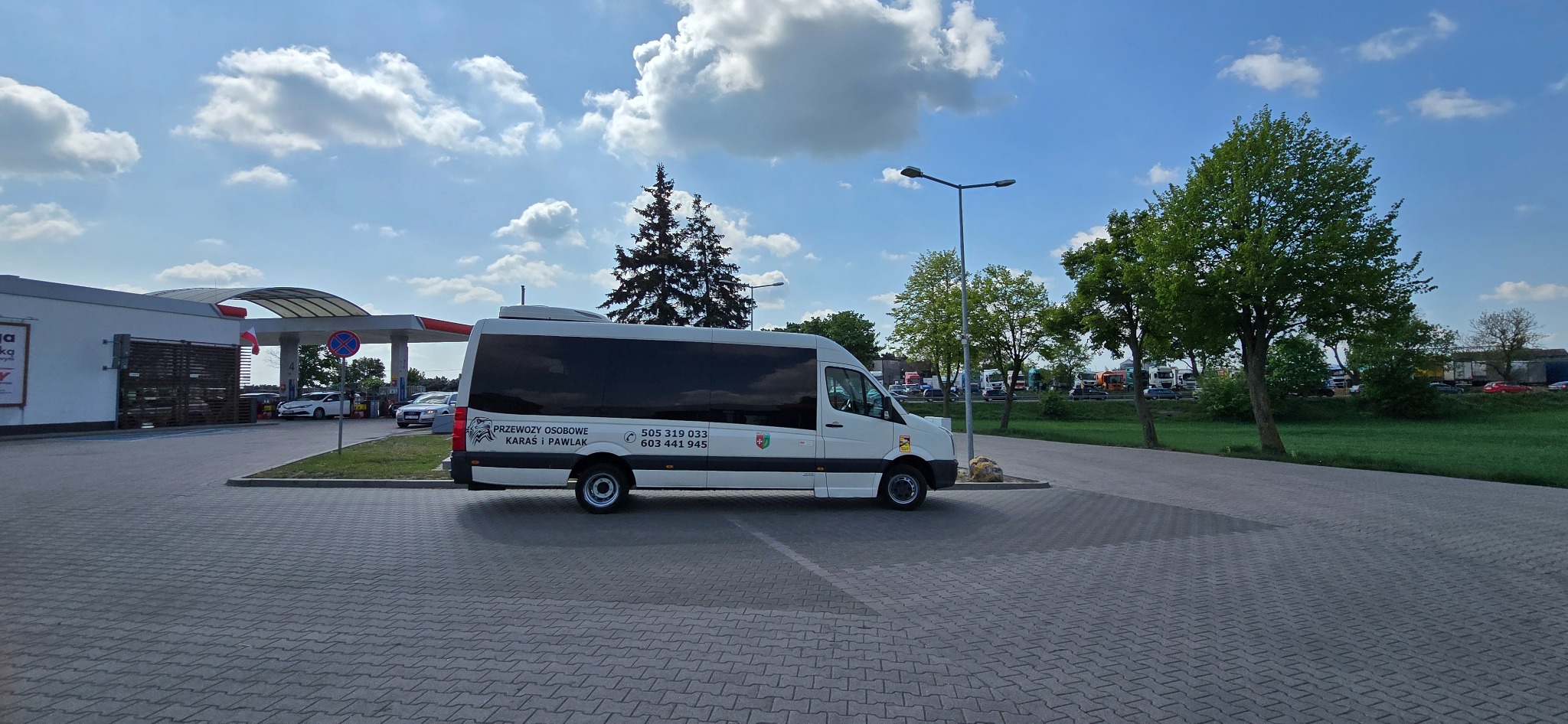 Biały bus z przyciemnianymi szybami, z logo firmy 'Przewozy Osobowe Karaś i Pawlak' zaparkowany na parkingu stacji benzynowej w słoneczny dzień.