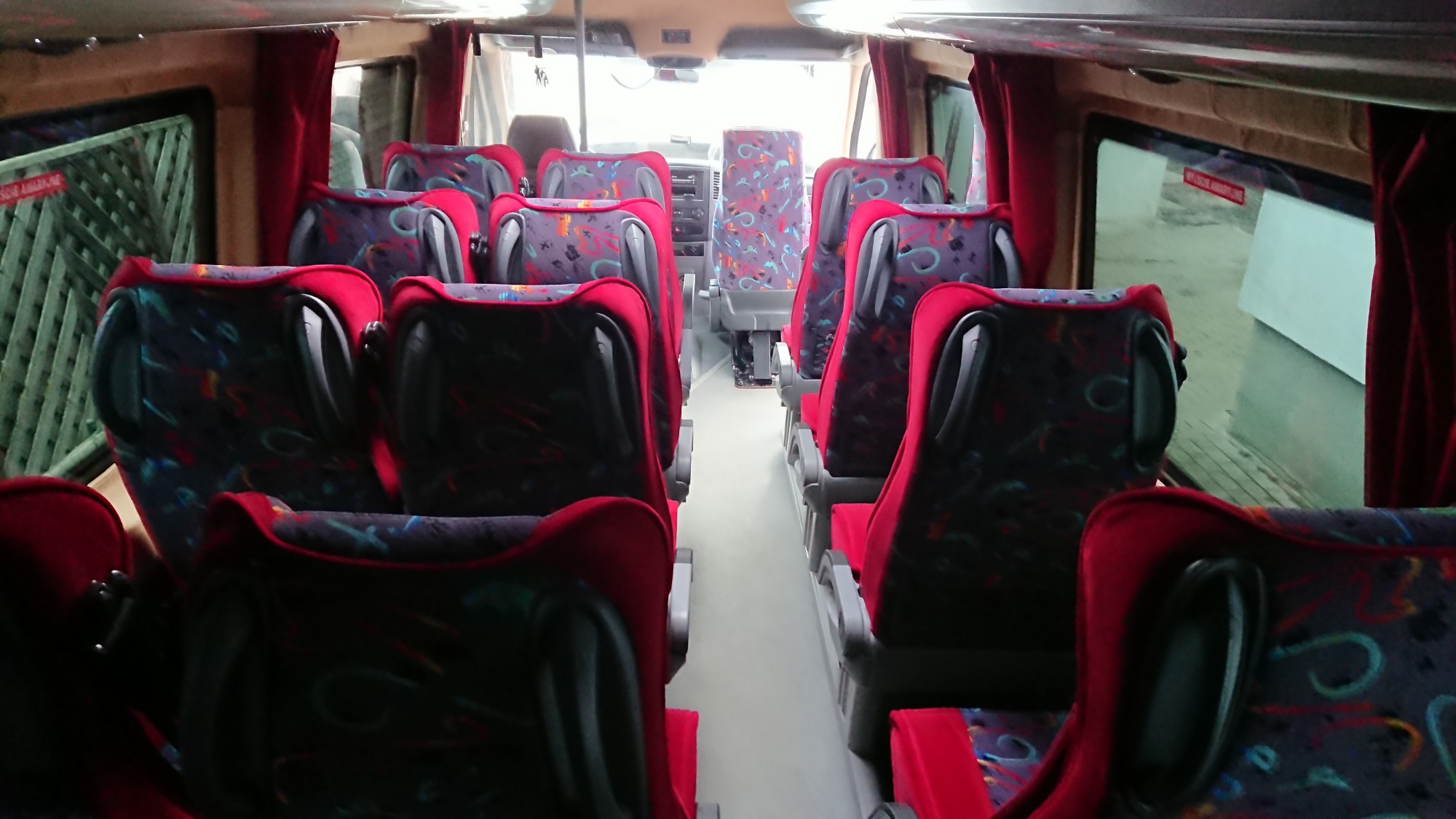 Wnętrze autobusu z czerwonymi zasłonami i fotelami o wzorzystej tapicerce, widok z tyłu pojazdu.