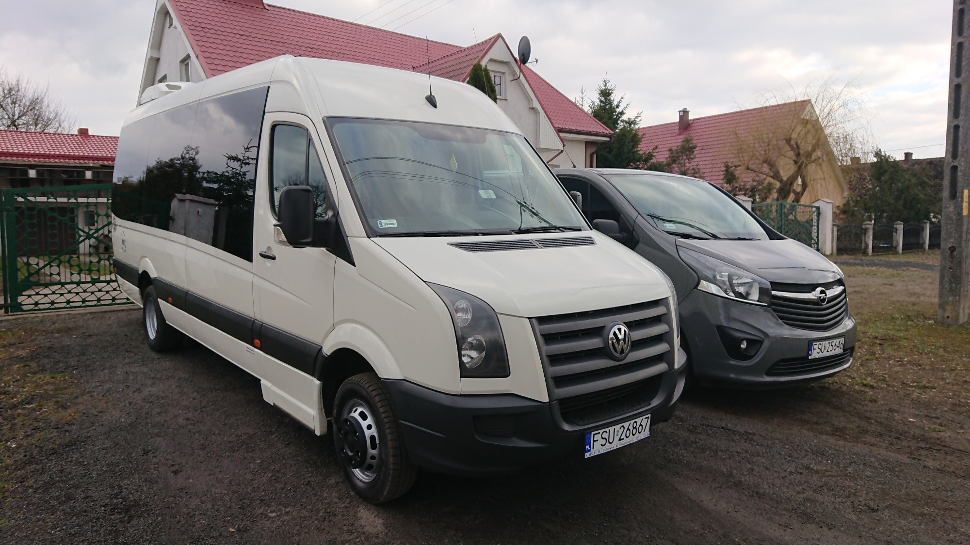 Biały bus Volkswagen Crafter z przyciemnianymi szybami i czarnymi felgami, zaparkowany obok szarego Opla Vivaro na szutrowej drodze przed domem z czerwonym dachem.