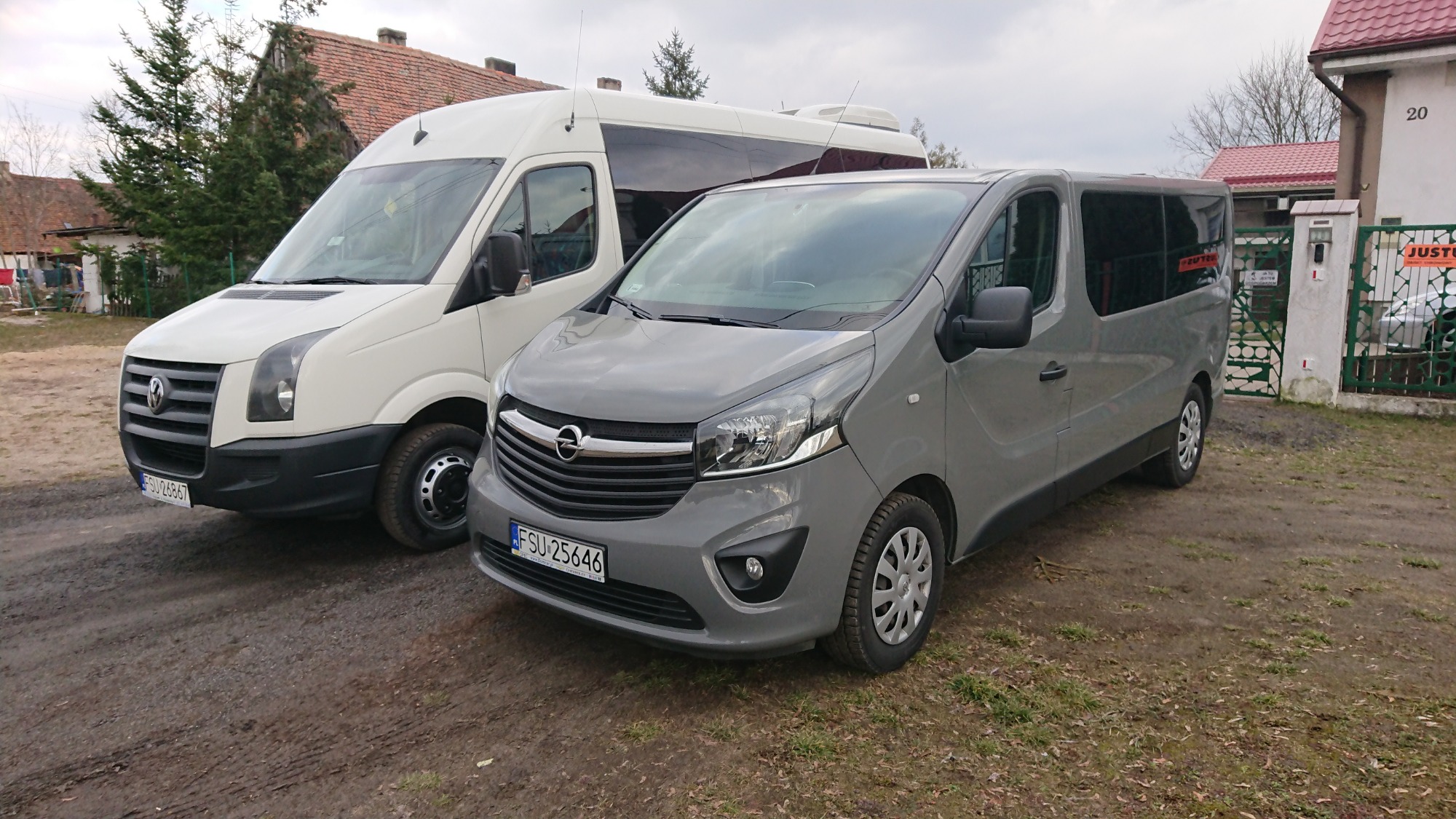 Dwa busy: biały Volkswagen Crafter i szary Opel Vivaro stojące obok siebie na błotnistym terenie przed domem z numerem 20 i zieloną bramą.