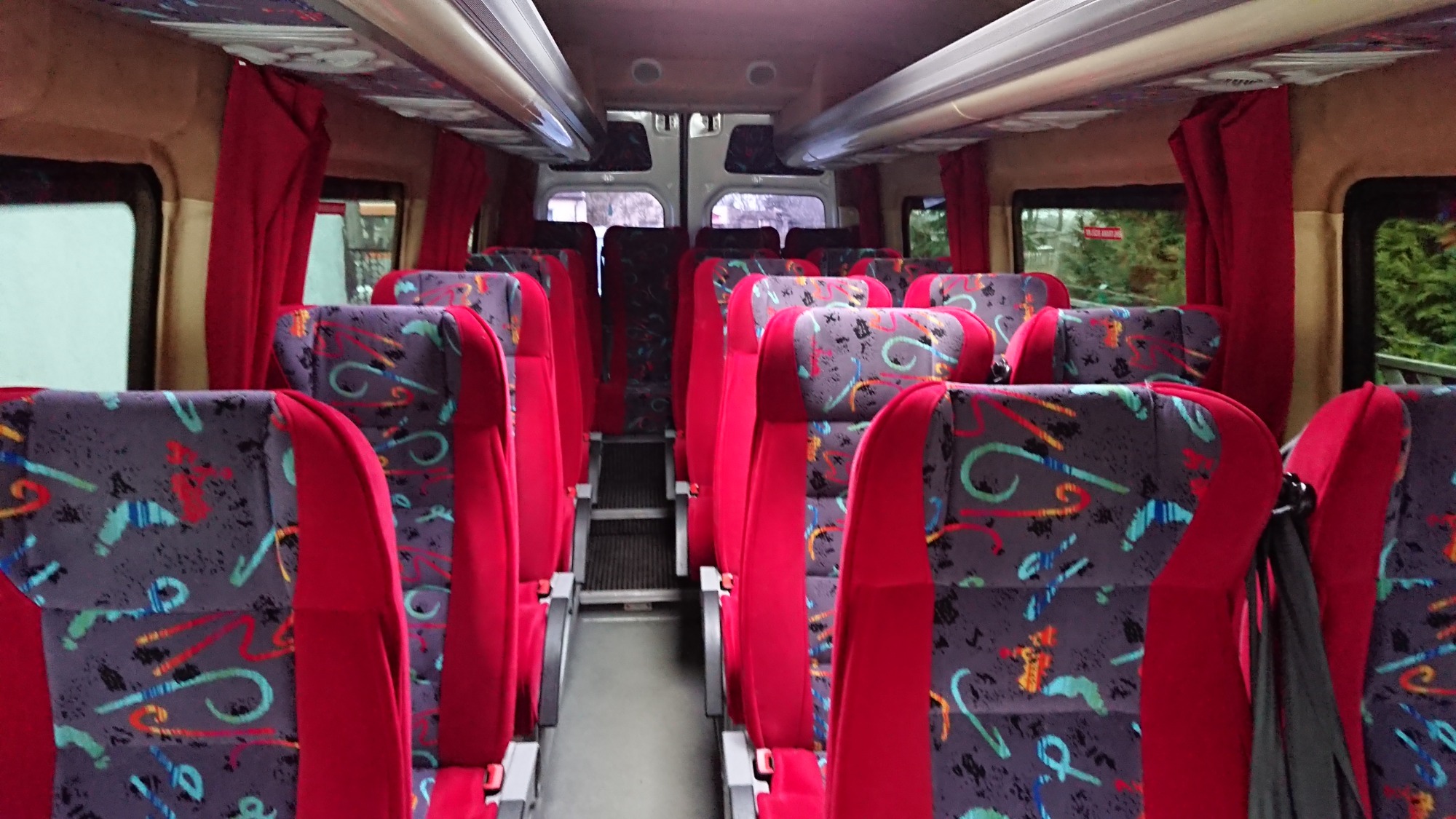 Wnętrze autobusu z czerwonymi fotelami o wzorzystych zagłówkach, zasłonięte okna z widokiem na zieleń.