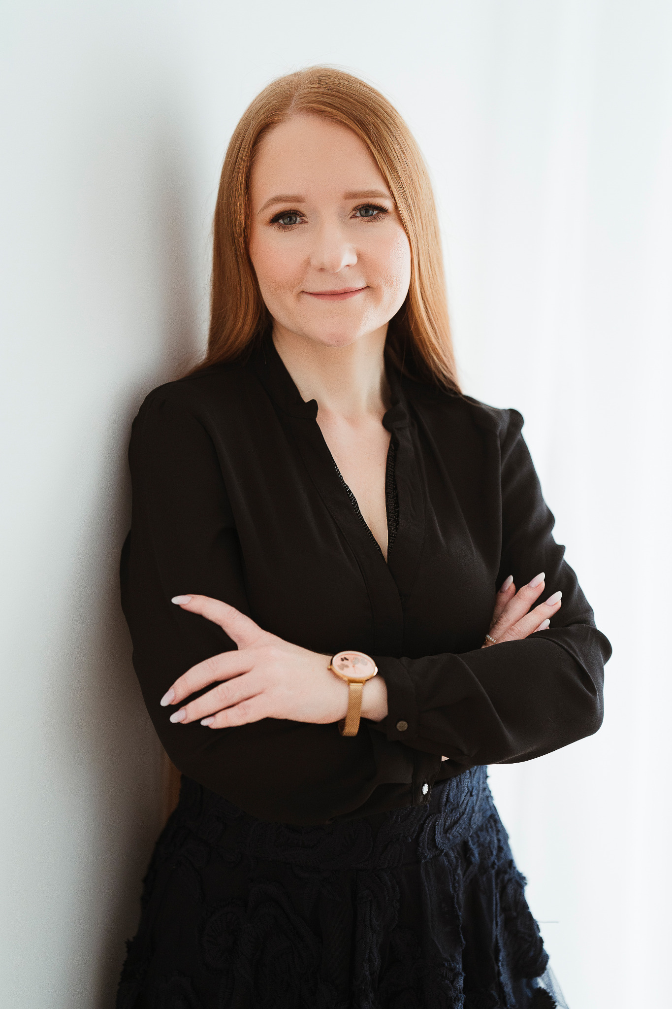 Portret kobiety z długimi rudymi włosami, ubranej w czarną bluzkę i ciemną spódnicę, stojącej na tle białej ściany z założonymi rękami i zegarkiem na nadgarstku.