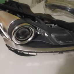 Reflektor samochodowy z elementami chromowanymi, widoczne ślady demontażu i uszkodzenia obudowy, leżący na białym tle.