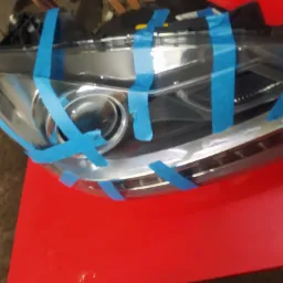 Samochodowa lampa przednia oklejona niebieską taśmą maskującą, przygotowana do renowacji lub naprawy, leżąca na czerwonym stole.