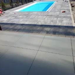 Wykończona powierzchnia wokół basenu z szarych, prostokątnych płyt betonowych, widoczne narzędzia i rękawice robocze, w tle ogrodzenie z prefabrykatów betonowych.