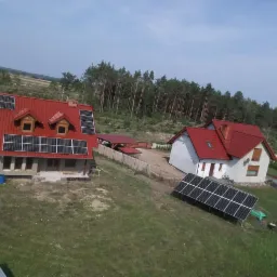 Dwa domy jednorodzinne z panelami słonecznymi na dachach i na ziemi, widok z góry, w tle las i łąki.