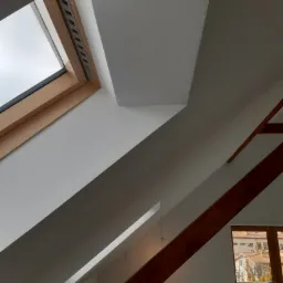 Poddasze po remoncie z oknem dachowym, świeżo pomalowane na biało, widoczne drewniane elementy konstrukcyjne i grzejnik pod oknem.