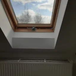 Poddasze z oknem dachowym z widokiem na pochmurne niebo i drzewa, poniżej biały grzejnik panelowy.