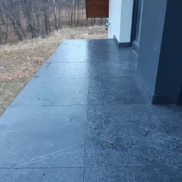 Wyłożony ciemnymi płytkami taras z widoczną fakturą kamienia, w tle jesienny krajobraz z drzewami bez liści.