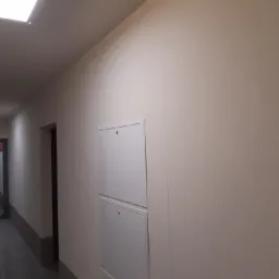 Pomieszczenie po remoncie, ściany pomalowane na jasny kolor, widoczne dwie białe szafki wbudowane w ścianę, w tle korytarz z widocznymi narzędziami malarskimi.