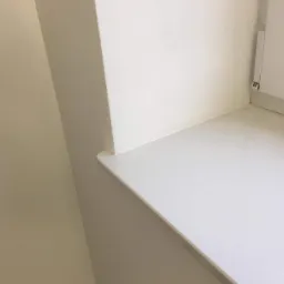 Fragment wnętrza po remoncie: gładka, beżowa ściana i biały parapet z oknem, widoczne łączenie ściany i parapetu.