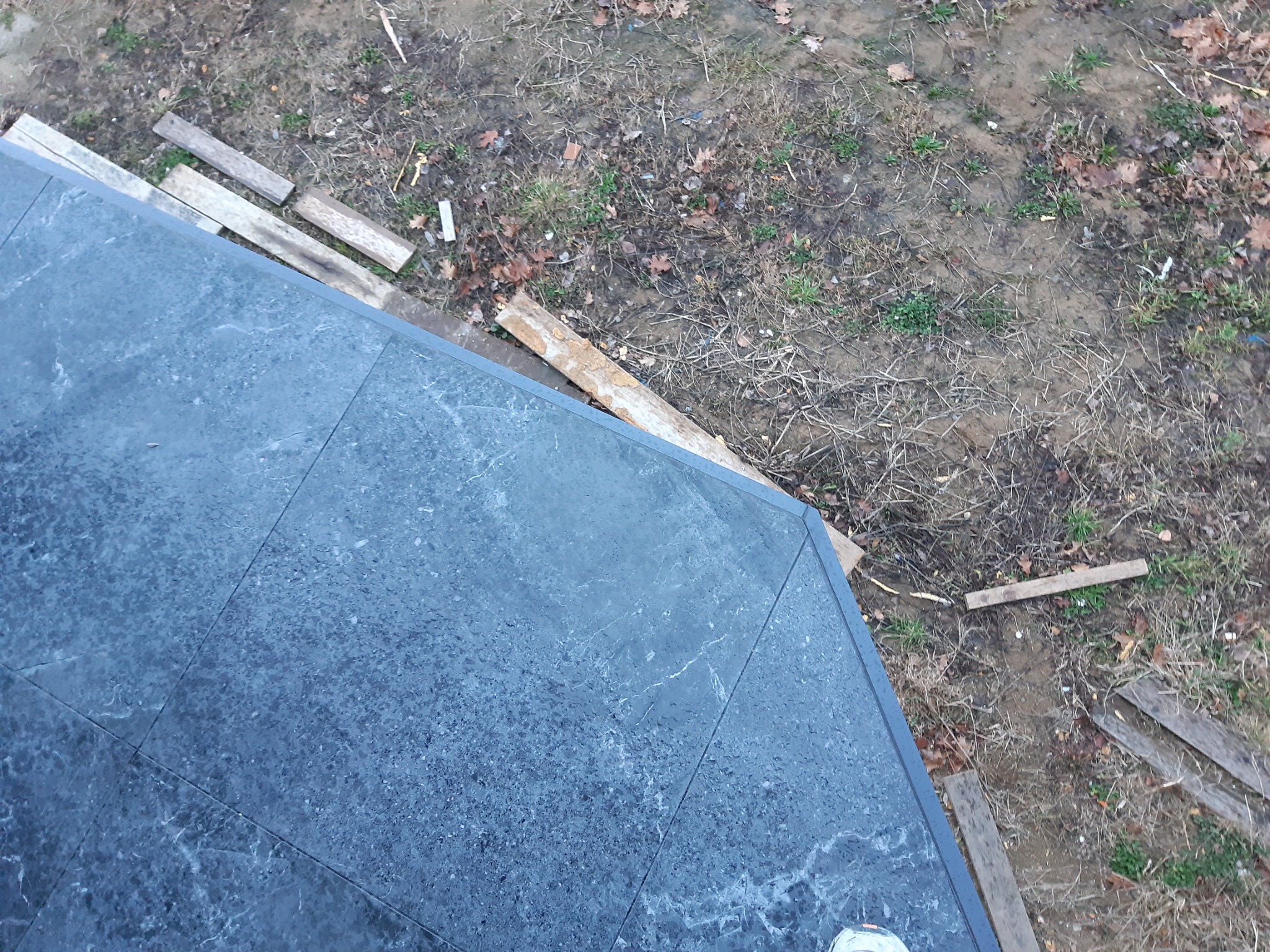 Widok z góry na fragment szarej płyty karton gips, ułożonej na zewnątrz, obok leżą drewniane deski i ziemia z liśćmi.