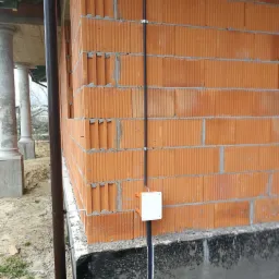 Czarny przewód elektryczny przymocowany do ściany z czerwonej cegły ceramicznej, z białą puszką instalacyjną na dole, widoczne elementy orynnowania i fundamenty budynku w trakcie budowy.