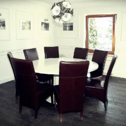 Sala konferencyjna z okrągłym stołem i ośmioma krzesłami z brązowej skóry, oświetlona nowoczesną lampą wiszącą, z obrazami na ścianach w białych ramach.