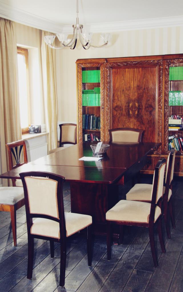 Sala konferencyjna z dużym drewnianym stołem i krzesłami obitymi kremowym materiałem, w tle regał z książkami i dekoracyjna lampa sufitowa.