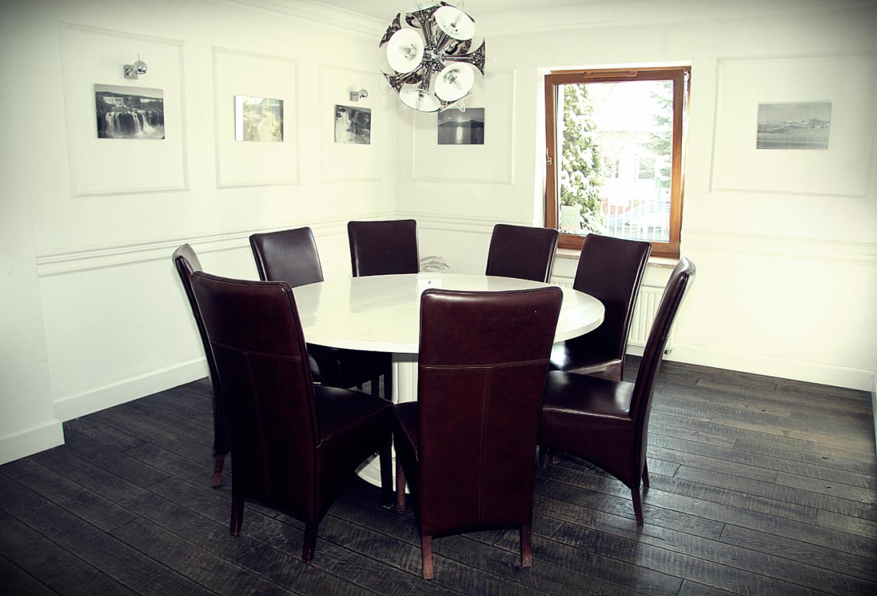 Sala konferencyjna z okrągłym stołem i ośmioma krzesłami z brązowej skóry, oświetlona nowoczesną lampą wiszącą, z obrazami na ścianach w białych ramach.