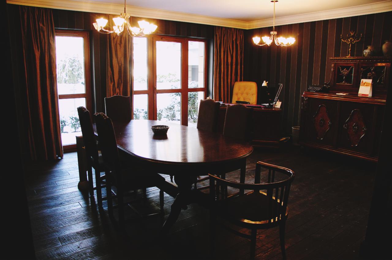 Wnętrze gabinetu z dużym, owalnym stołem z ciemnego drewna, krzesłami, biurkiem i kredensem w stylu retro, oświetlone lampami w stylu art deco, z widokiem na zimowy krajobraz za oknem.