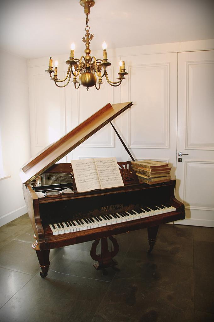 Antyczny fortepian marki Ant. Petrof z otwartą klapą, nutami i stosem książek, oświetlony przez złoty żyrandol w klasycznym wnętrzu z białymi panelami ściennymi.