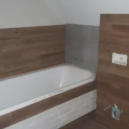 Biała wanna w łazience z drewnopodobnymi panelami ściennymi i białymi płytkami, widoczne przyłącza wodne i częściowo zakryta folią ochronną.