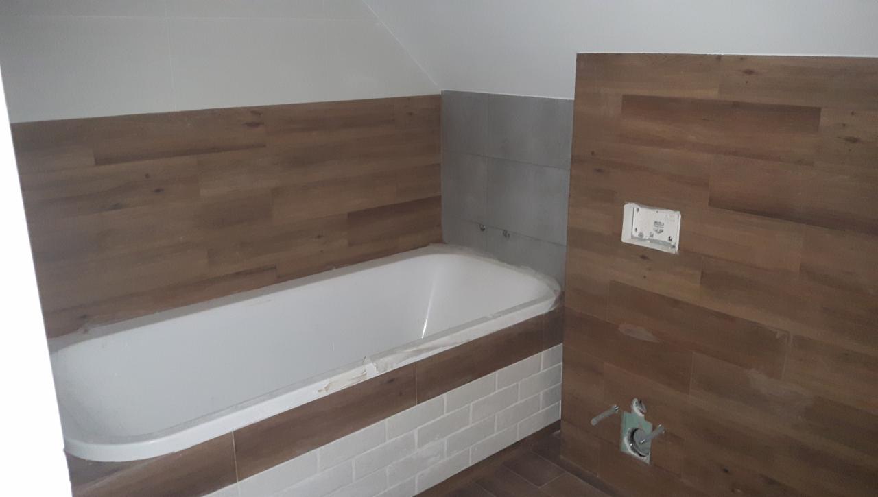 Biała wanna w łazience z drewnopodobnymi panelami ściennymi i białymi płytkami, widoczne przyłącza wodne i częściowo zakryta folią ochronną.