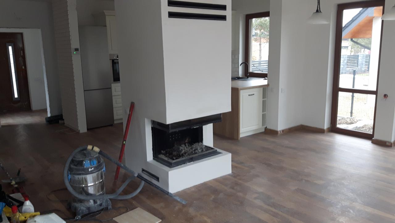 Wnętrze domu w trakcie wykańczania, z widocznym kominkiem obudowanym na biało, częściowo ułożonym parkietem, odkurzaczem przemysłowym i poziomnicą opartą o kominek.