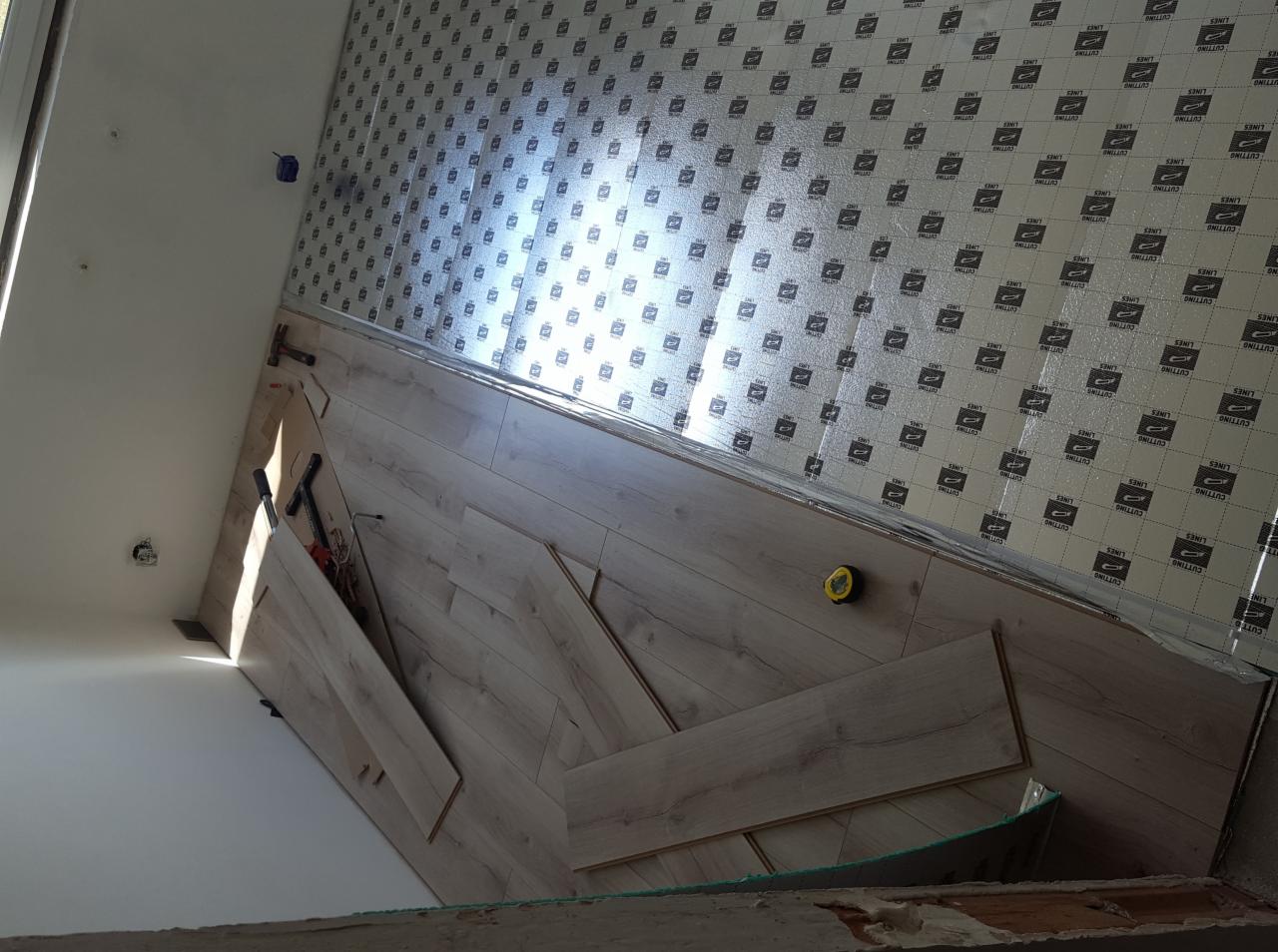 Układanie jasnego parkietu laminowanego na podkładzie izolacyjnym, widoczne narzędzia: młotek, miarka zwijana i przymiar kątowy.