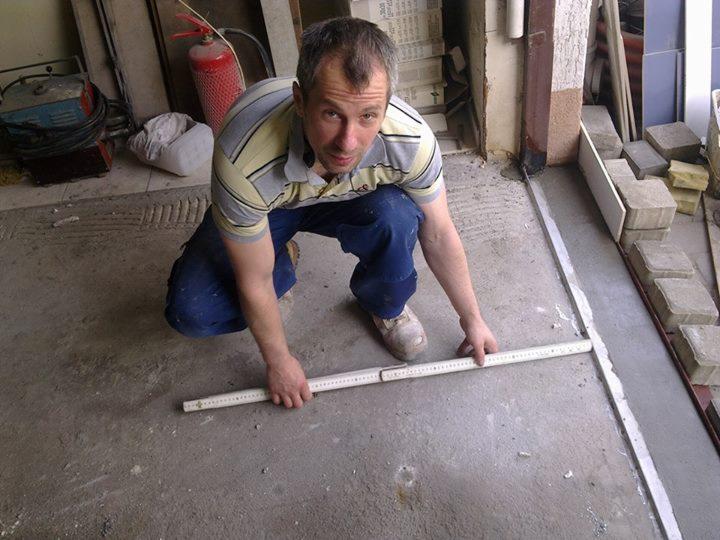 Mężczyzna w kucki mierzy długość posadzki przed ułożeniem kostki brukowej, widoczny fragment ściany z niebieskimi płytkami i gaśnica w tle.