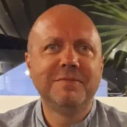 Portret mężczyzny w średnim wieku, z krótkimi włosami i lekkim zarostem, uśmiechniętego, na rozmytym tle z elementami roślinności i budynku.