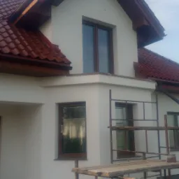 Dom jednorodzinny z nową, jasną elewacją i brązową stolarką okienną, częściowo zasłonięty przez metalowe rusztowanie, widoczne schody wejściowe.