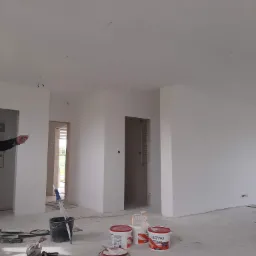 Wnętrze w trakcie malowania: świeżo pomalowane na biało ściany, widoczne puszki z farbą Cieszynka, narzędzia malarskie i wiadro na podłodze, przygotowane do dalszych prac wykończeniowych.