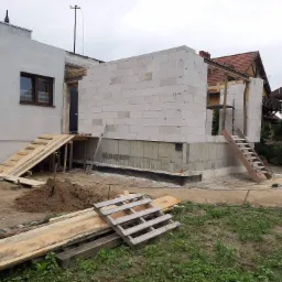 Budowa domu jednorodzinnego: widok na wznoszone ściany z bloczków betonowych, widoczne fundamenty z izolacją, drewniane rampy i schody, oraz stos desek na ziemi.