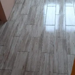 Ułożone pod kątem płytki podłogowe imitujące drewno w pomieszczeniu z grzejnikiem i fragmentem ściany pomalowanej na różowo; widoczna gąbka i śrubokręt na podłodze.