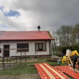 Montaż czerwonej blachodachówki na dachu domu jednorodzinnego; jeden pracownik na dachu, drugi przycina blachę na ziemi, widoczne rusztowanie i przygotowane materiały.