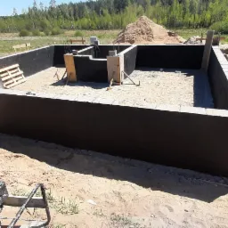 Wykopane fundamenty domu jednorodzinnego z izolacją pionową ścian, widoczny ubijak wibracyjny, paleta i stos styropianu w tle.