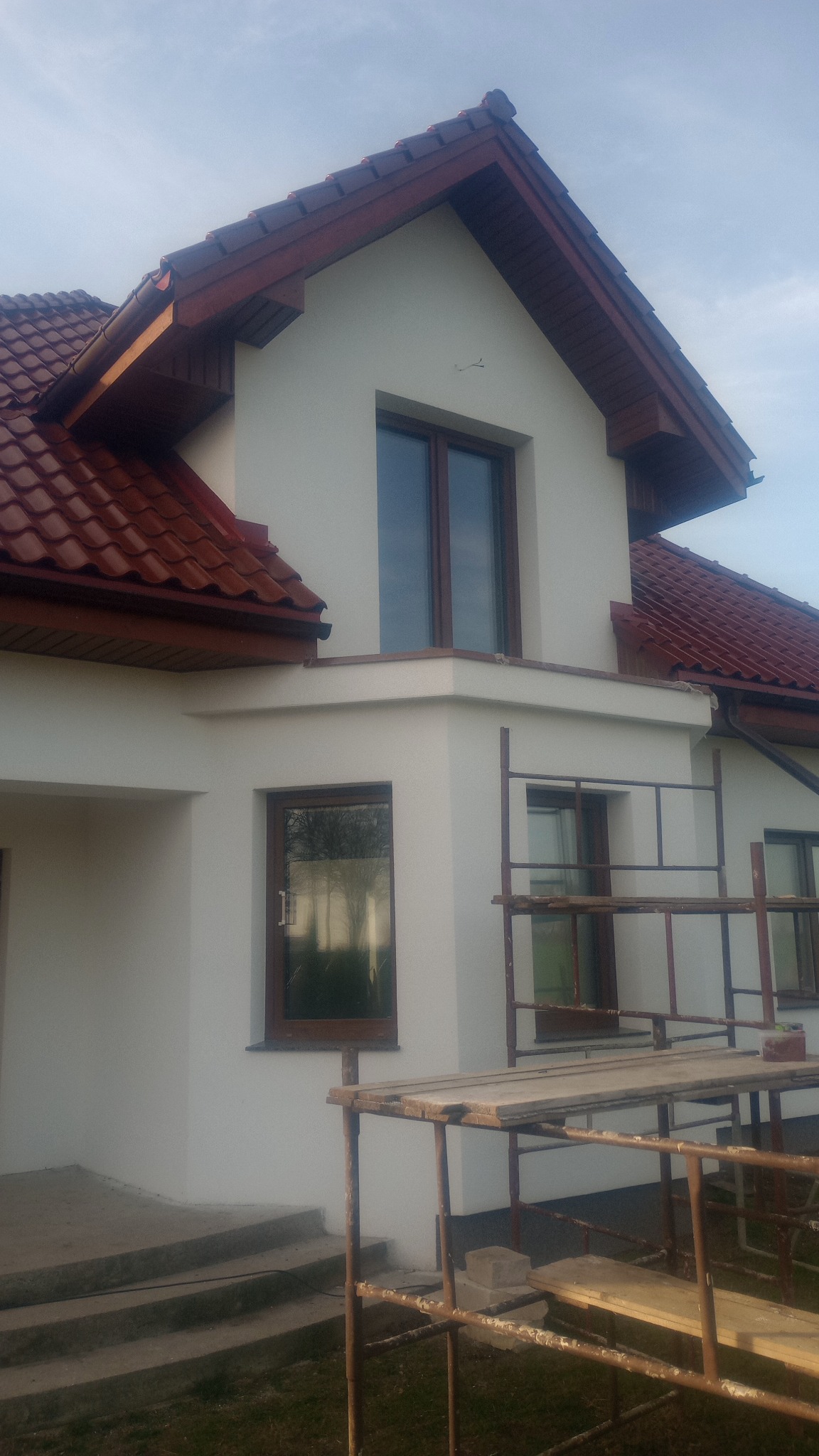 Dom jednorodzinny z nową, jasną elewacją i brązową stolarką okienną, częściowo zasłonięty przez metalowe rusztowanie, widoczne schody wejściowe.