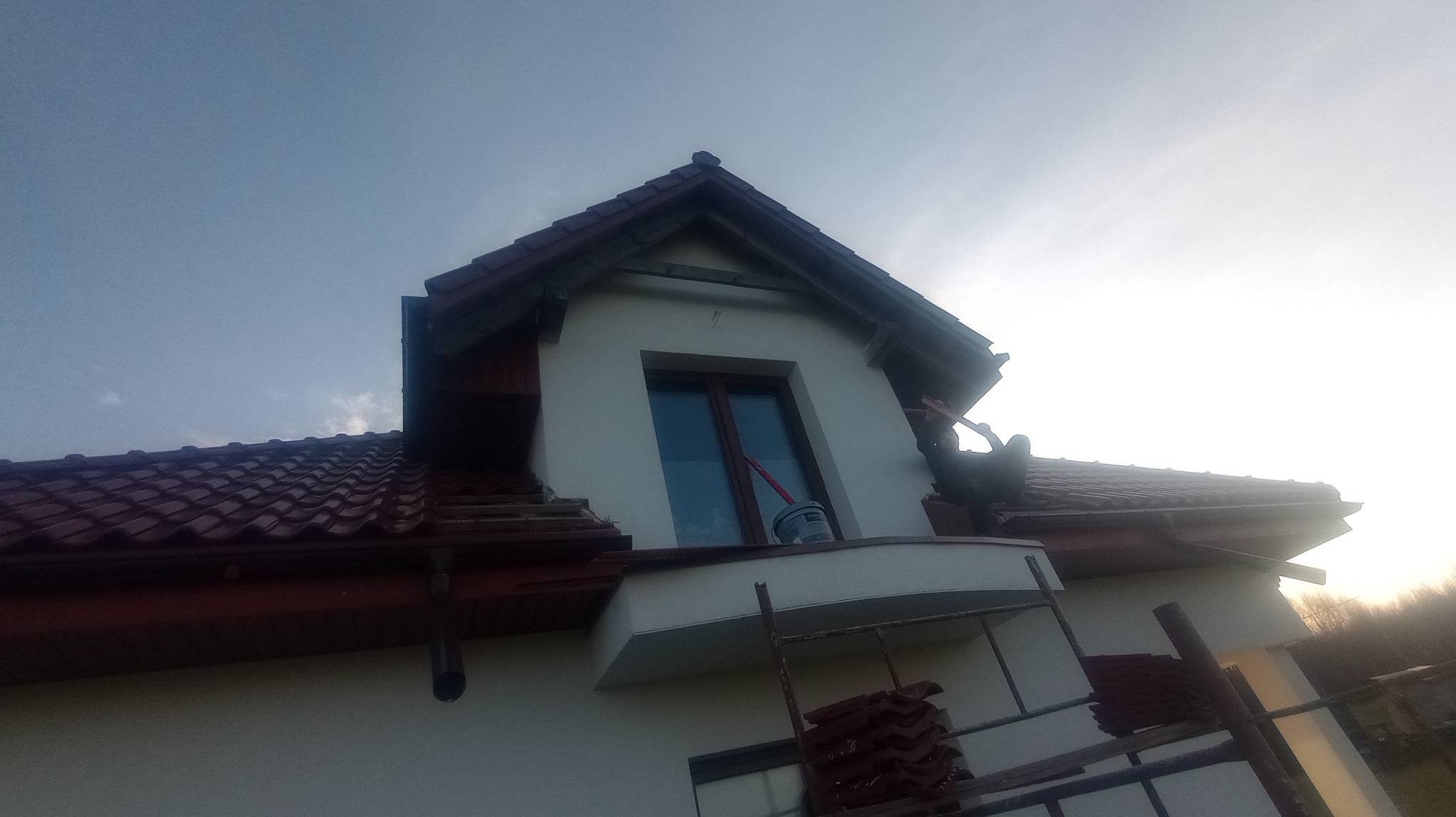 Prace wykończeniowe na elewacji domu: malowanie fragmentu ściany przy oknie, widoczne rusztowanie, wiadro z farbą i pracownik w trakcie pracy.