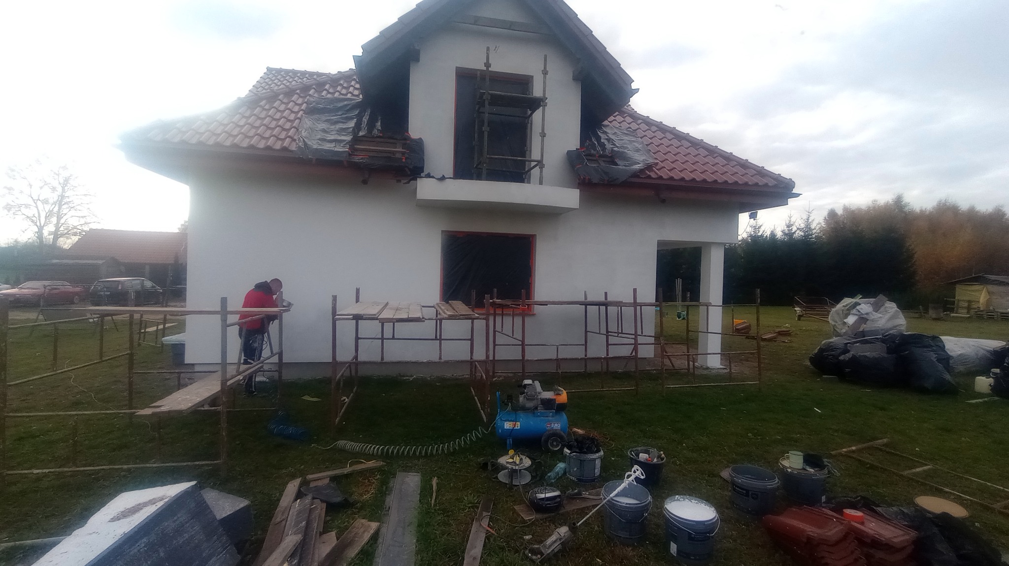 Praca na elewacji domu jednorodzinnego: mężczyzna w czerwonej kurtce pracuje na rusztowaniu przy białej ścianie, widoczne narzędzia, wiaderka z farbą i kompresor, okna zabezpieczone folią, dach...