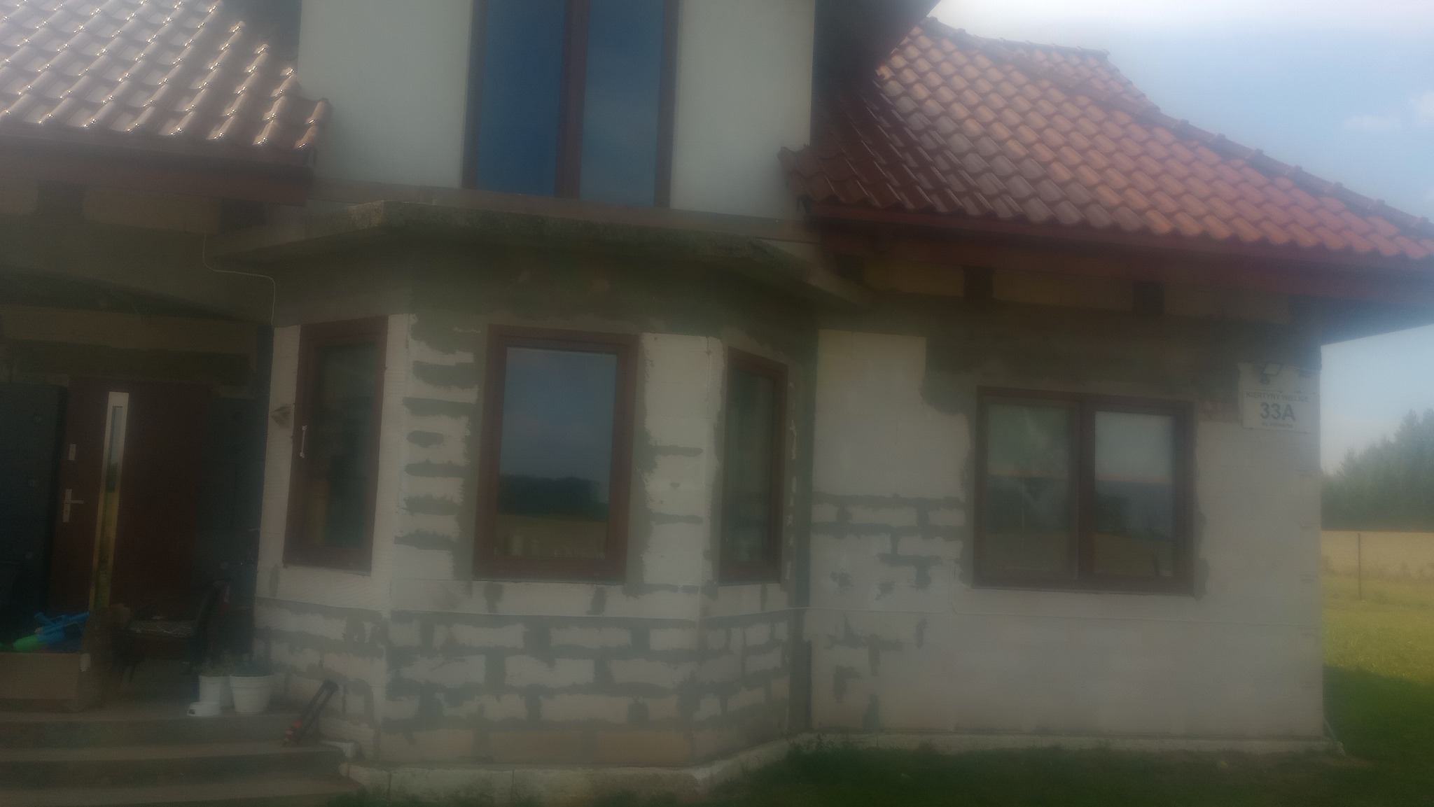 Dom jednorodzinny w trakcie ocieplania elewacji, widoczne bloczki i częściowo nałożony klej, brązowe ramy okienne, dach z czerwonej dachówki, tabliczka z numerem posesji 33A.