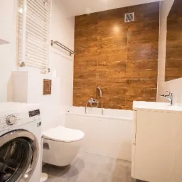 Nowoczesna łazienka z drewnopodobnymi płytkami na ścianie, białą armaturą i pralką Electrolux po lewej stronie.