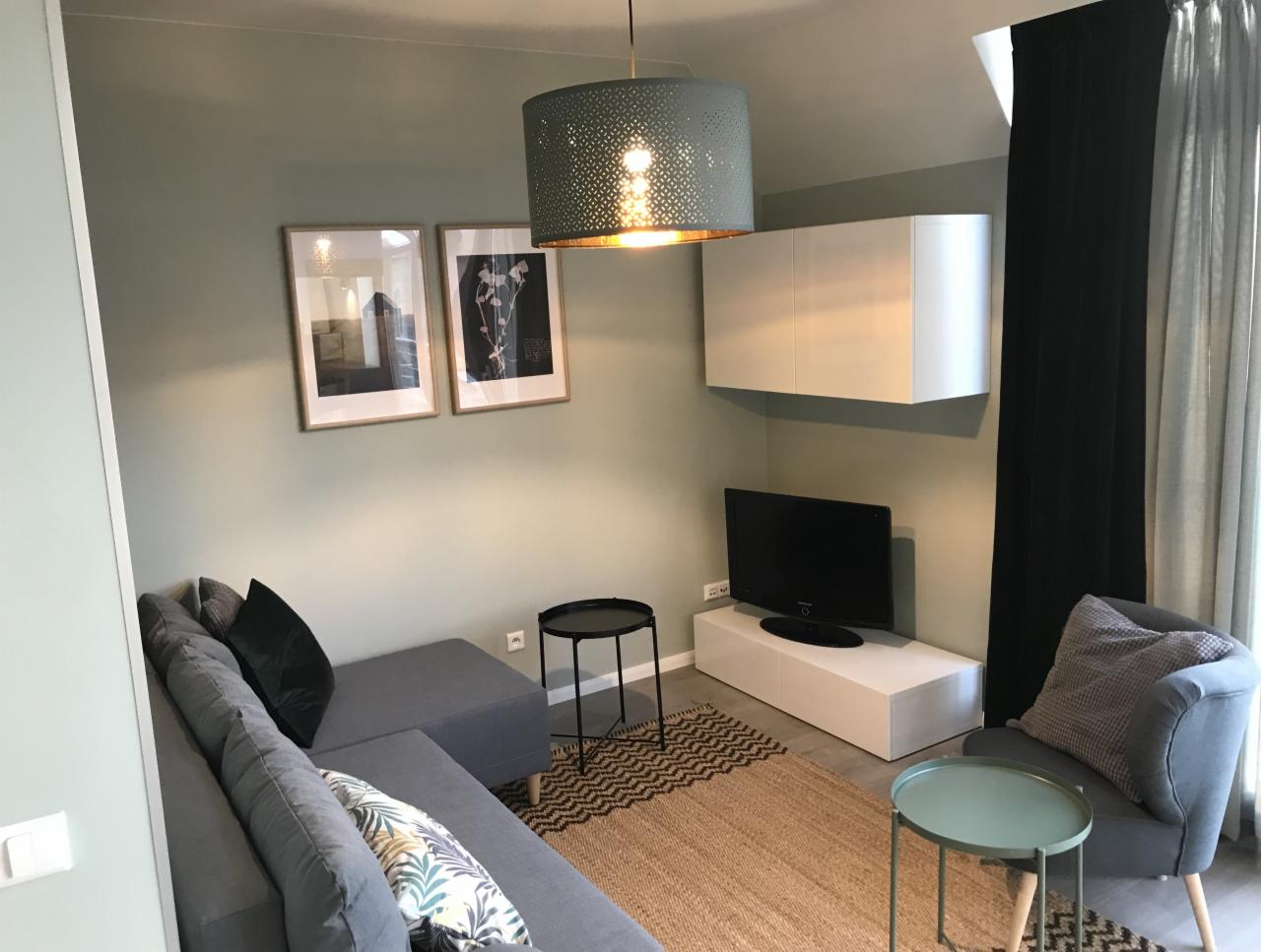 Nowoczesny salon z szarą sofą narożną, dywanem w geometryczny wzór i wiszącą szafką, oświetlony ażurową lampą. Dwa obrazy w ramkach wiszą na ścianie w kolorze szałwii.