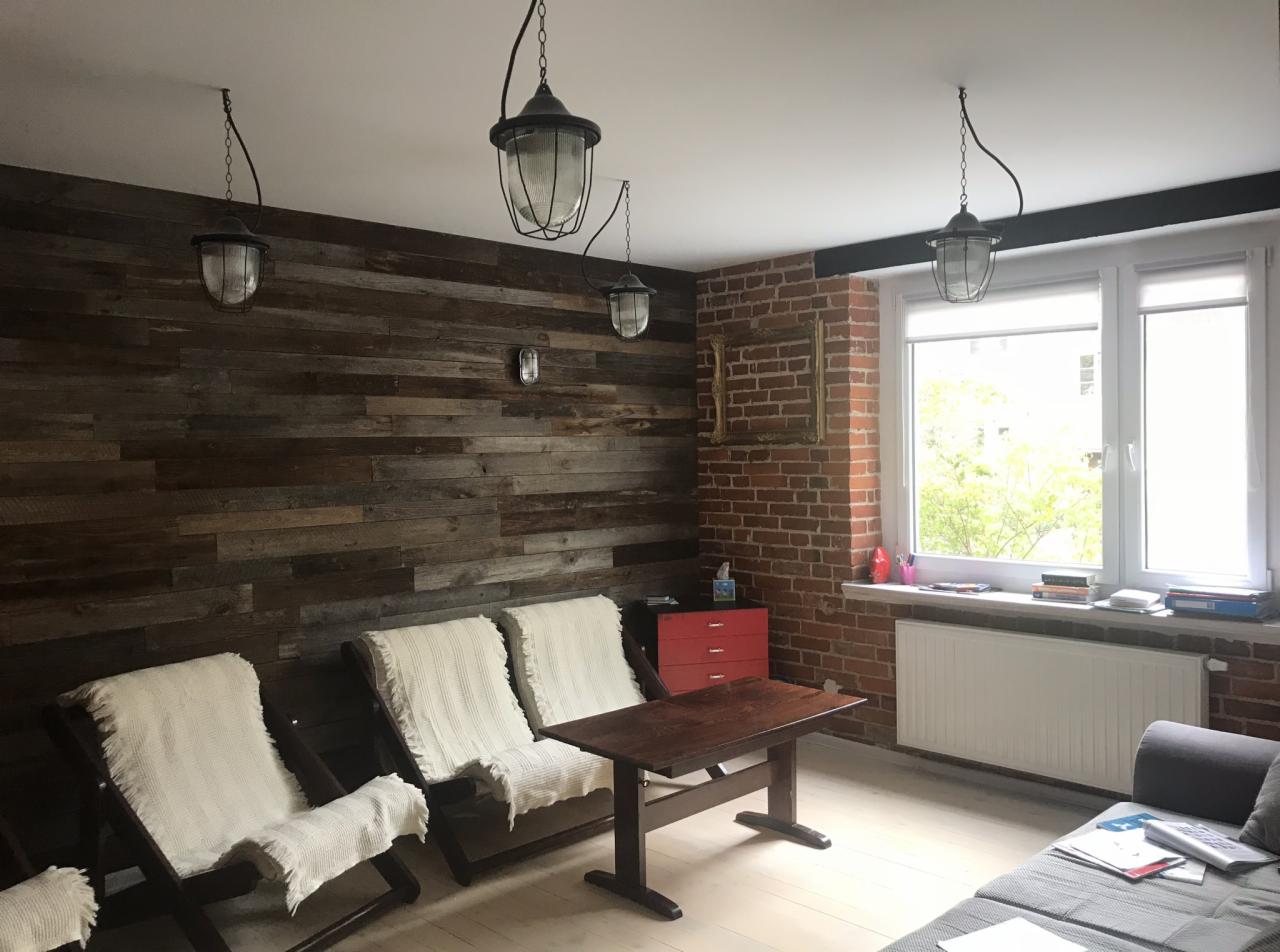 Salon z drewnianą ścianą z desek, trzema lampami wiszącymi w stylu industrialnym i ceglanym filarem okiennym z dekoracyjną ramą, uzupełniony trzema leżakami z białymi narzutami.