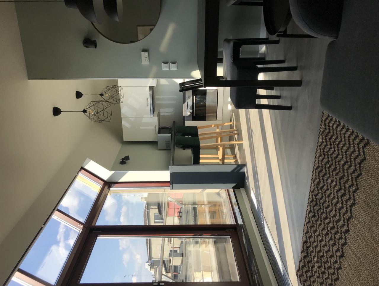 Jasne wnętrze mieszkania z dużym oknem dachowym, widok na kuchnię z drewnianymi stołkami barowymi, nowoczesne lampy wiszące i szary dywan z geometrycznym wzorem.