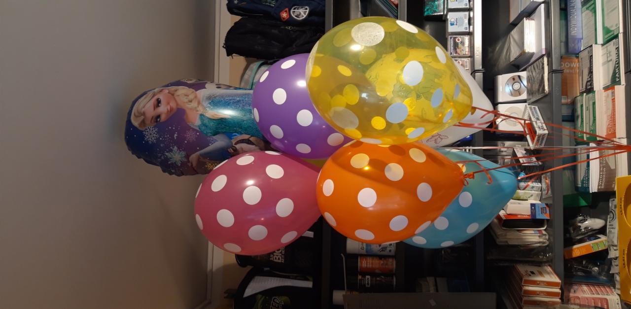 Kolorowe balony z motywem Elsy i w kropki, unoszące się w pomieszczeniu z regałem pełnym przedmiotów w tle. Dekoracja urodzinowa lub imprezowa.